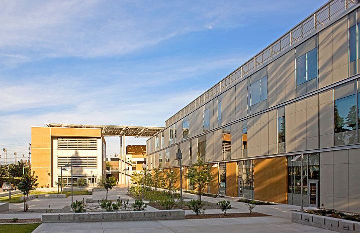 Image 1. A 3-story modern building clad with beige and terra cotta-colored panels. The building wraps around a minimalist outdoor plaza on the left side.