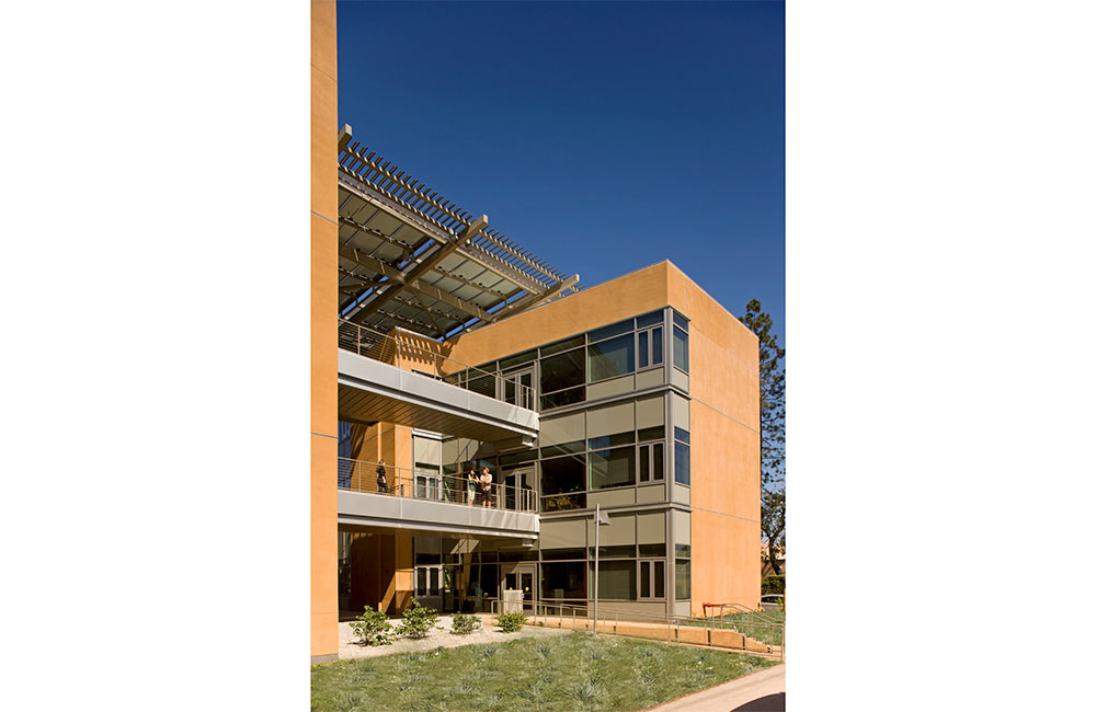 Image 3. Corner view of a 3-story terra cotta-colored modern building with large glass windows.  Silver metal pedestrian walkways extend towards the left.