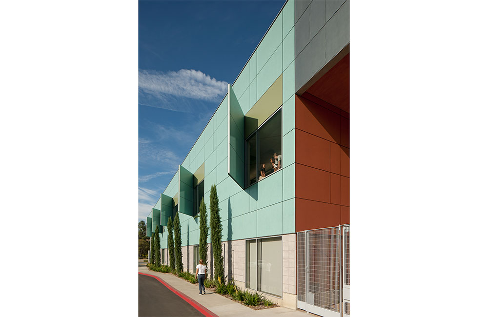 Image 2. Side view of 2-story building showing turquoise-colored panels on upper level and concrete colored tiles on the first floor. Vertical sunshade panels pivot outwardly from the second-floor windows. 