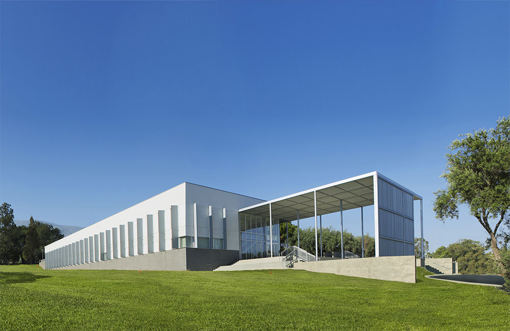 Image 1. Corner exterior view of linear white 2-story modern building with angled vertical sunshade panels on the left facade and a large slatted entry canopy that turns into a vertical slat screen wall on the right side.
