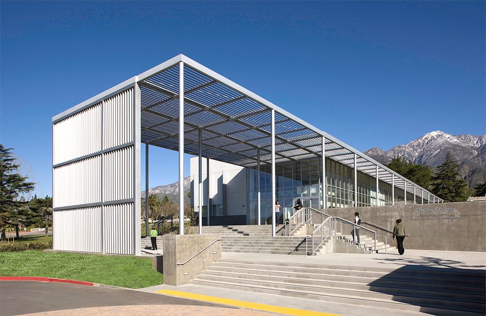 Image 2. Corner exterior view of building entry with a large slatted canopy above that turns into a vertical slat screen wall.  A series of steps and platforms seen in the foreground lead to the front entrance.