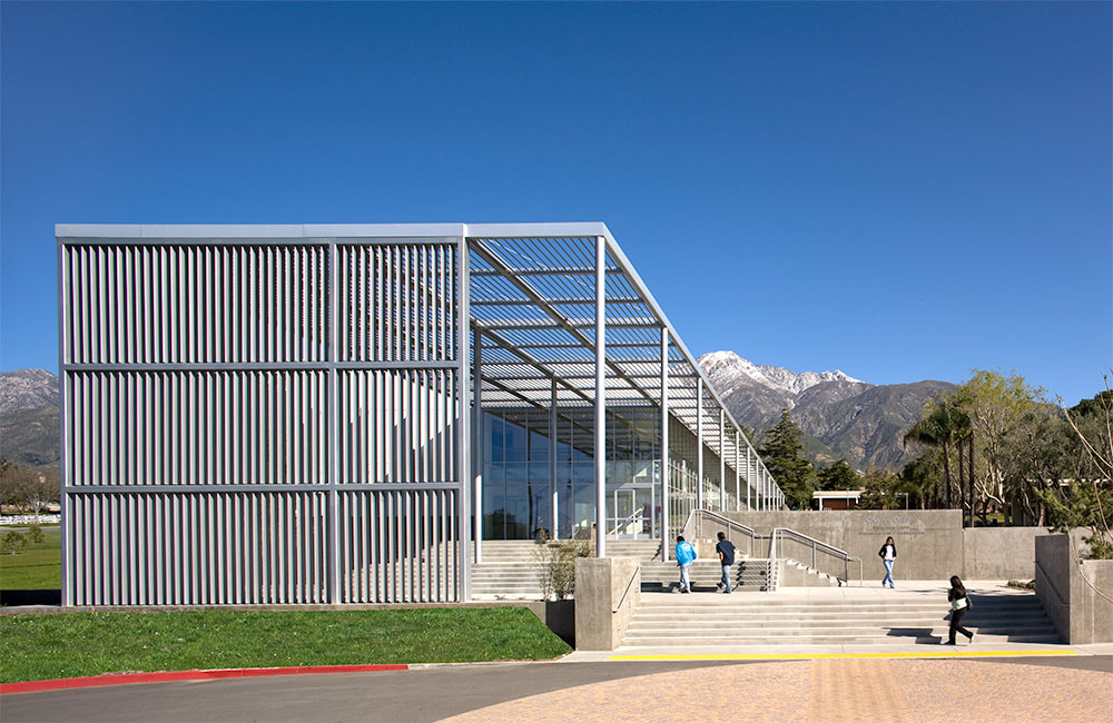 Image 6. Front exterior view of building entry with a large slatted canopy above which turns into a vertical slat screen wall. A series of steps and platforms seen in the foreground lead to the front entrance.